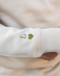 Primer plano del bordado de inicial y corazón verde caqui en la manga de la sudadera beige