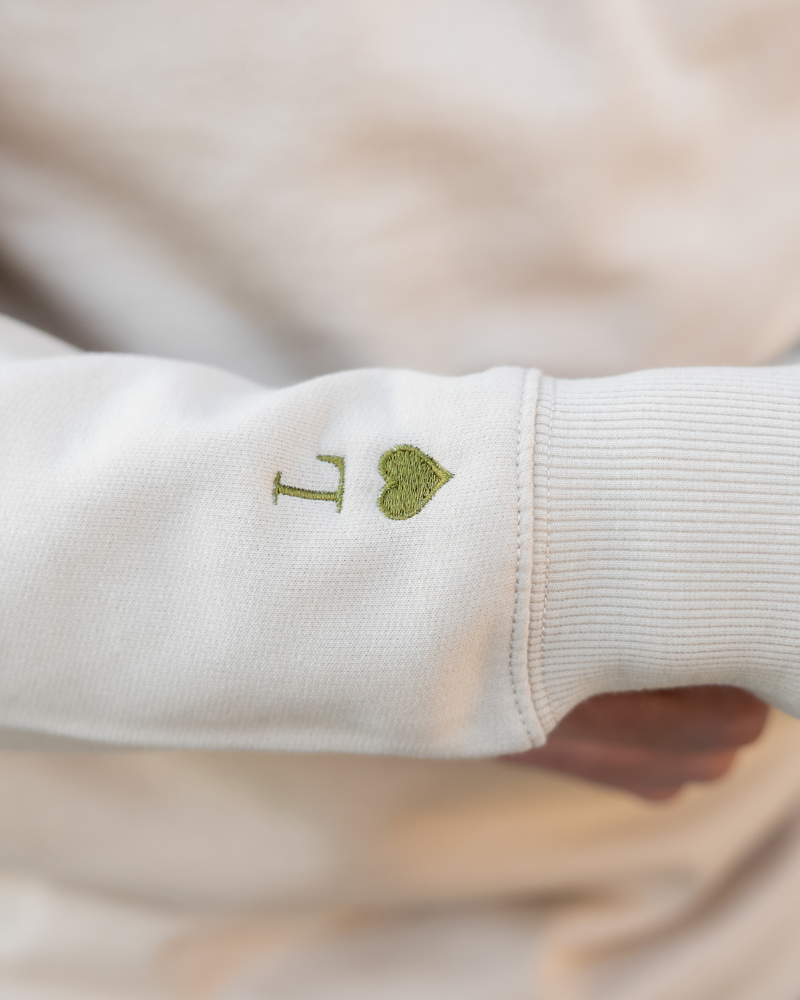 Primer plano del bordado de inicial y corazón verde caqui en la manga de la sudadera beige
