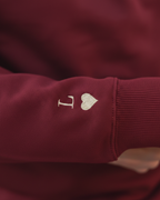 Primer plano del bordado de inicial y corazón granate en la manga de la sudadera granate